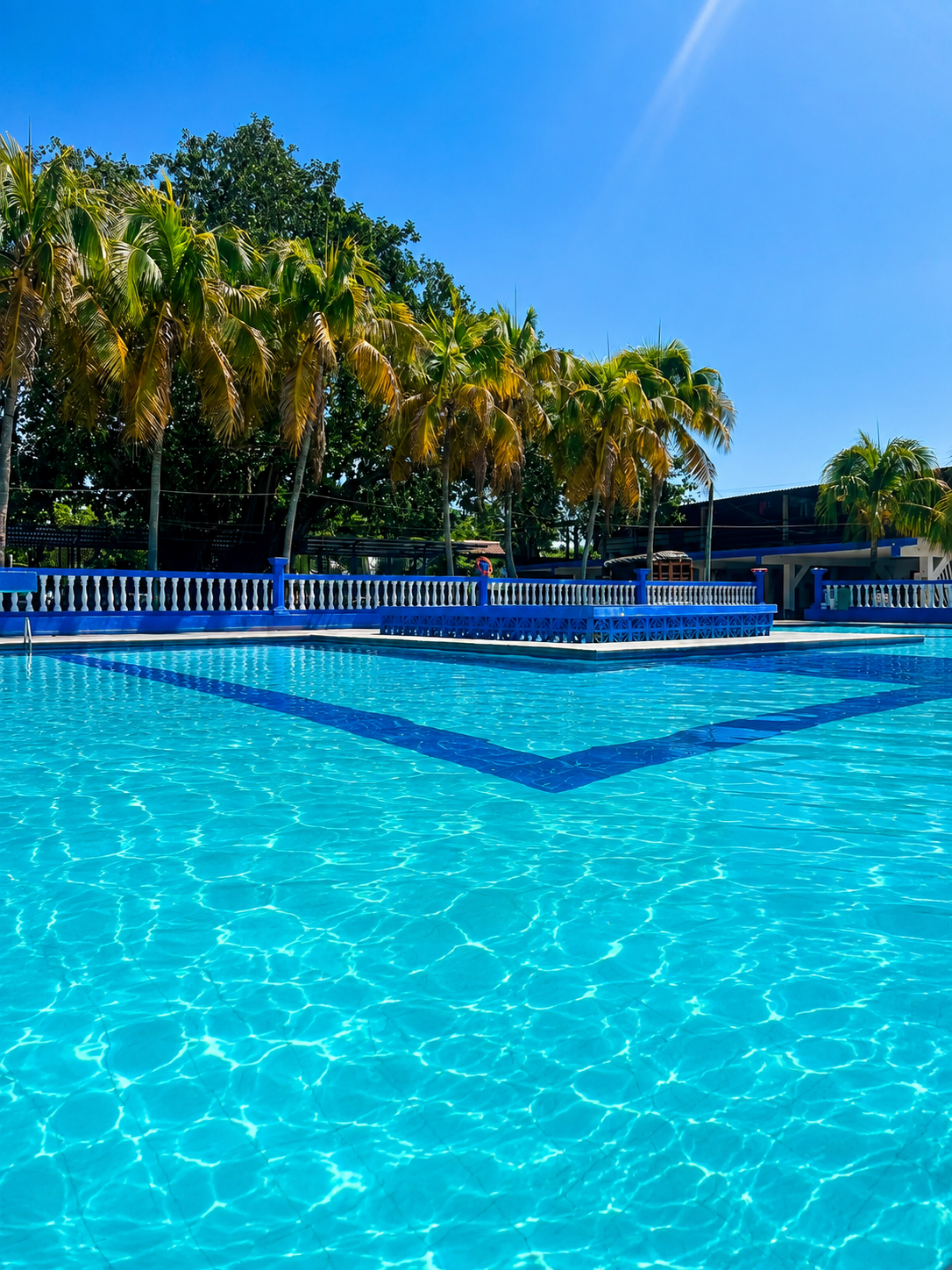 Piscina del Hotel Zona Tropical en La Dorada, Caldas: agua turquesa cristalina, palmeras tropicales y barandas azules bajo un cielo despejado. Disfrútala con tu habitación sencilla.